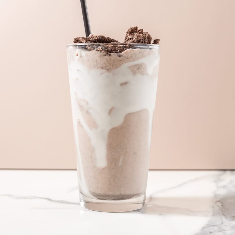 A tall glass of chocolate milkshake with whipped cream and cookie crumbles, on a marble counter.