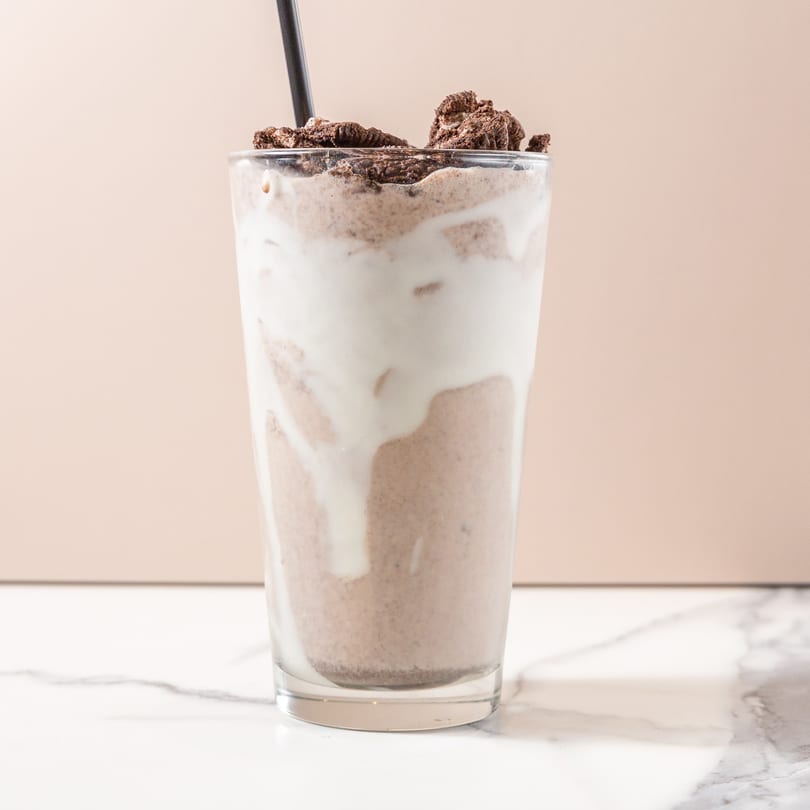 A tall glass of chocolate milkshake with whipped cream and cookie crumbles on a marble surface.