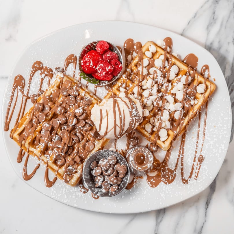 Two waffles with chocolate drizzle, chocolate chips, white chocolate chips, ice cream, and berries.