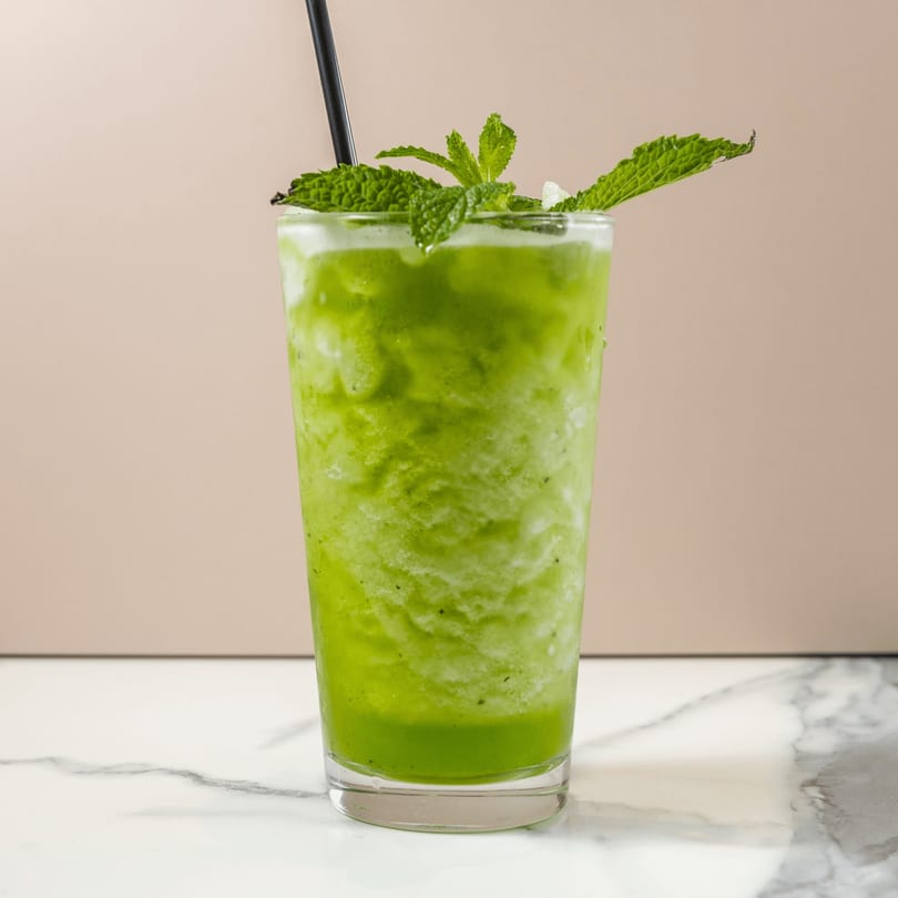 A vibrant green smoothie with mint leaves and a black straw, on a marble surface.