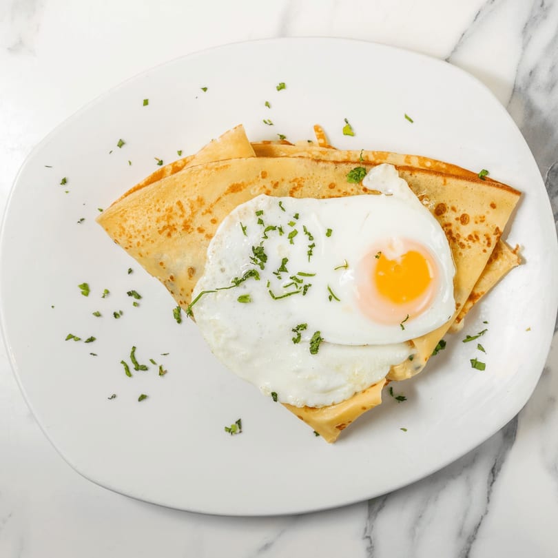 Savory crepe topped with a fried egg, melted cheese, and fresh herbs on a white plate.