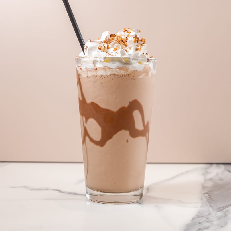 Chocolate milkshake with whipped cream, nuts, and a black straw on a marble counter.