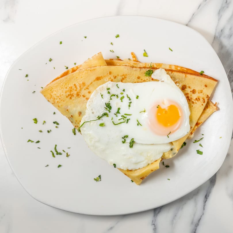 Savory crepe topped with a fried egg, melted cheese, and fresh parsley on a white plate.