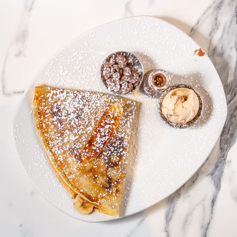A banana crepe dusted with powdered sugar, served with chocolate chips, caramel, and ice cream.